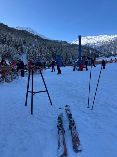 This image displays an outdoor, snow-covered alpine ski resort scene under a clear blue sky. While the prompt specifies Anger, Austria, the visual evidence strongly suggests a high-alpine ski environment with extensive mountain ranges and infrastructure, which is not characteristic of Anger, Styria.

In the immediate foreground, two orange skis with black bindings, one labeled "Metron 10," lie flat on the snow. To their right, two black ski poles are planted vertically. A black, three-legged high-top table with a red circular top is positioned in the center-left foreground.

The midground features approximately 15-20 individuals, all dressed in diverse, brightly colored winter ski apparel including jackets, pants, and helmets. Many are standing on skis, suggesting active participation or recent activity. A person in a blue jacket stands prominently in the center-right midground. To the left of the high-top table, several red and wooden lounge chairs are arranged in the snow, occupied by a few individuals. In the mid-right, a tall, prominent blue cylindrical pillar stands upright, possibly part of a ski lift system.

The background is dominated by densely forested mountainsides, heavily laden with snow, and extending upwards to higher, snow-capped peaks. A ski lodge or building structure with a visible deck is situated in the far right background. The overall scene depicts a busy recreational area at a ski resort.