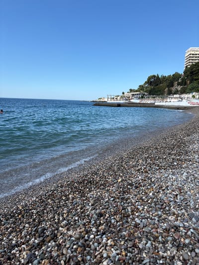 The image depicts a tranquil pebble beach scene under a clear blue sky. The foreground is dominated by a vast expanse of multi-colored pebbles, wet near the waterline where gentle waves lap the shore. In the mid-ground, the deep blue sea stretches out, with a few swimmers visible enjoying the water. To the right, a rocky coastline rises, covered in lush green trees, and a white building with many balconies is perched on the hillside. A white structure, possibly a pier or promenade, extends into the sea, with some wooden walkways and white canopies visible. A red sign with white lettering is positioned on this structure. The overall atmosphere is one of a peaceful summer day at a seaside resort.