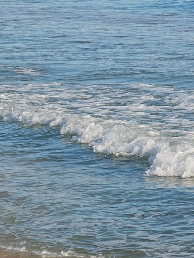 The image shows a close-up of the ocean at Deveses, Spain, on a sunny day. The water is a clear blue, with gentle waves rolling towards the shore. White foam is visible on the crest of the waves. The background shows more calm water, with ripples reflecting the sunlight. There are no people or other objects visible in the scene. The environment is the natural seascape of the Mediterranean Sea.