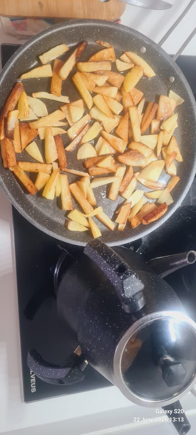 In Dalaman, Turkey, a close-up, top-down shot captures french fries being fried in a pan. The french fries are golden brown and appear to be cut into irregular shapes and sizes. They are spread across the non-stick surface of the pan, some with oil glistening on them. The pan itself is dark gray with a speckled texture, and its handle is visible on the right side. Below the pan, a black electric stovetop is visible, and a black kettle with a speckled finish and a silver rim is positioned on the stovetop, partially obscuring the lower portion of the pan and its contents. A wooden cutting board and a knife are also visible in the upper portion of the image, out of focus. The overall impression is of a domestic kitchen setting where food is being prepared.