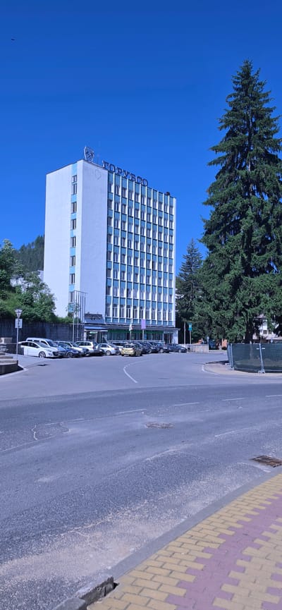 The image shows a tall, white, and blue hotel building with the Cyrillic inscription "ГОРУБСО" on its roof, likely indicating the hotel's name and location in the city of Madan, Bulgaria. The building is multi-story with numerous windows. In front of the hotel, there is a parking lot filled with several cars. A large evergreen tree stands to the right of the hotel, casting a shadow. The foreground shows a paved road and sidewalk, with some construction barriers visible on the right side of the image. The sky is clear and blue, suggesting a sunny day. There are no people visible in the frame.