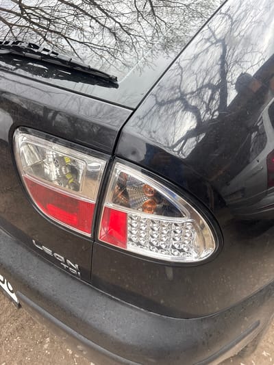 The image shows the rear of a black Citroen C3 car, likely photographed in Debnevo, Bulgaria. The car's right tail light is visible, composed of a white, orange, and red lens. The license plate is partially visible at the bottom. The car appears to be parked on pavement near what looks like dirt or gravel. Tree branches are reflected on the rear window. The car's paint is dusty and worn.