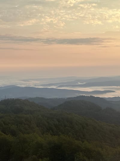 This is a landscape photograph taken from a high vantage point overlooking a valley filled with rolling hills and mountains. The sky is a soft gradient of pink and pale yellow, with scattered clouds. In the middle ground, a layer of fog blankets the lower portions of the mountains, creating a sense of depth and distance. The foreground is dominated by dark green, densely forested hills. The overall impression is serene and tranquil, suggesting a natural setting, possibly at sunrise or sunset. There are no people or animals visible in the image. The photograph appears to depict a rural or mountainous region. The location is specified as Beli Osam, Bulgaria, which is known for its natural beauty and mountainous terrain.