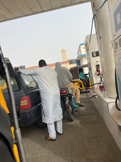 This image depicts a fuel station in Basawa, Nigeria, under a hazy, overcast sky. In the immediate foreground, the left side of a dark blue sedan or hatchback is visible, positioned at a fuel pump. A male individual, wearing a white traditional garment (dankali) and brown sandals, stands next to the car's rear, facing away from the viewer. Behind him, a second male, dressed in a gray hooded sweatshirt and khaki pants, sits astride a dark-colored motorcycle, also facing away from the viewer. Further back in the queue, a yellow motorized tricycle (keke napep) is visible, with an individual in a teal-colored garment at its controls. On the right, two fuel pumps are visible; the closer pump has a red fuel nozzle extracted and resting on its side, with a red bucket placed on the ground nearby. The pumps stand on a white tiled base, underneath a light-colored canopy structure. In the background, beyond the fuel station, a building with a blue and white facade is visible, along with another structure featuring a brown roof. Prominently in the distance, a tan-colored mosque minaret topped with a golden dome rises above the other buildings. The ground is concrete.