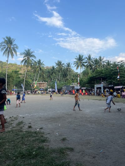 The image depicts an outdoor community gathering in Desa Batu Layar, Indonesia, under a clear blue sky with scattered white clouds. The scene is dominated by a large, open dirt field where multiple children are playing football. One child in a grey shirt and shorts actively dribbles a football in the lower right, while another in an orange shirt walks toward the center-left. Other children are visible across the field, with at least one near the center also engaging with a football.
The background features a dense line of tall palm trees and lush green hillside vegetation. Along the far edge of the field, numerous temporary structures, including stalls, tents, and a multi-level wooden/bamboo framework, are set up. These structures are adorned with various flags and colorful banners, including the red and white Indonesian national flag. A significant crowd of adults and children gathers around these background structures, suggesting a market, festival, or community event. The foreground consists of sparse grass and packed dirt with minor debris. The overall lighting indicates daylight conditions.