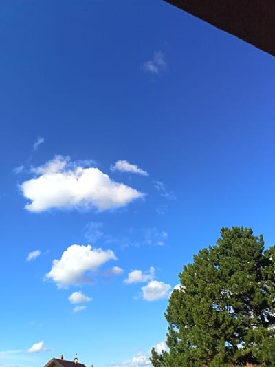 The scene depicts an outdoor environment in Klíčany, Czechia, under a clear, bright blue sky containing scattered white cumulus clouds of various sizes. In the lower right quadrant, the dense green foliage of a large coniferous tree occupies a significant portion of the frame. The upper portion of a building with an orange-brown tiled roof and a central reddish-brown chimney structure is partially visible at the bottom left. A dark, angular architectural element, possibly a roofline or beam, projects into the frame from the top right corner. No individuals or active subjects are present.