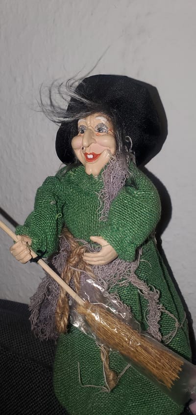 A doll resembling a witch is positioned against a white, textured wall. The doll wears a black hat and a green dress with a frayed, purple sash around its waist. It holds a small broom made of straw, enclosed in clear plastic. The doll's face is detailed with a prominent nose, smiling mouth with red lipstick, and glitter around the eyes. Its gray hair is windswept. The doll is placed on a dark, textured surface, possibly a sofa. There is no discernible connection to the city of Sasbach, Germany.