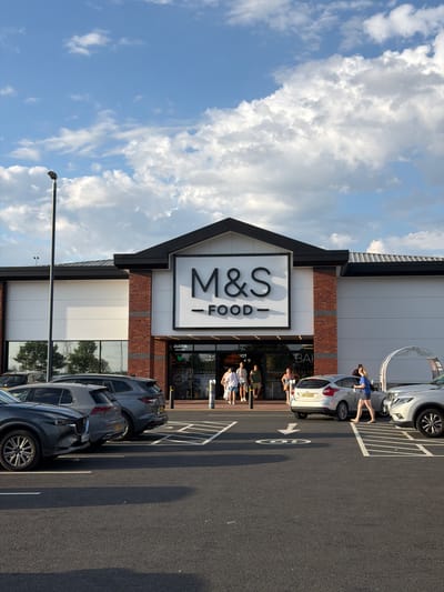 A M&S Food store in Cannock Chase, United Kingdom, is captured in this outdoor, eye-level shot on a cloudy day. The store's facade features prominent "M&S FOOD" signage in large black letters against a white background. Red brickwork accents the entrance area, which is flanked by large windows. Several vehicles are parked in the paved lot in front of the store, including a grey Audi Q5, a dark grey Mazda CX-5, a silver Ford Focus, and a silver SUV. People are seen walking into and out of the store. A tall black lamppost stands to the left of the frame. The sky is a mix of blue and white clouds.