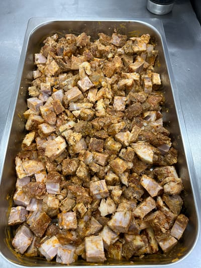 This is a top-down, medium shot of a rectangular metal pan filled with cubed, seasoned pork belly. The pork is cut into roughly one-inch cubes and appears to be raw or unmarinated, with visible layers of meat and fat. The seasoning is a mix of reddish-brown spices, some of which appear to be chili flakes, and possibly herbs, giving the pork a textured, slightly oily appearance. The pan is situated on a stainless steel countertop, likely in a commercial kitchen environment. The lighting is overhead and neutral. There are no people or other objects of significance in the frame. The background is dominated by the metallic surface of the countertop. The location is not specified in the image or context provided.
