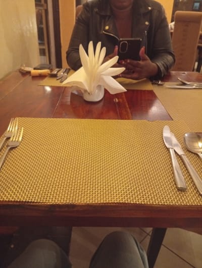 A restaurant table with a brown wood surface and a woven yellow placemat. Silverware, including a fork, knife, and spoon, are placed on the placemat. A napkin holder containing white napkins sits on the table. In the background, a person in a black leather jacket uses a mobile phone. The bottom of the image shows a person's legs in grey pants.