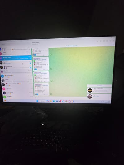 The image shows a computer monitor displaying a Telegram application window. The Telegram window is filled with chat messages, primarily related to financial transactions. Several messages show "tolovlar buypin" which likely translates to "payments buy" or "payment purchase." These messages contain amounts in UZS (Uzbekistani Som) and refer to card payments, PEYNET, and HUMO Card. Some messages also include dates, such as "20.06.2026." The top of the window shows the title bar with a search icon, settings icon, and other typical window controls, as well as "Перевод (русский)" indicating a Russian translation feature. The application appears to be running on a Windows operating system, as indicated by the Windows taskbar at the bottom of the screen, which includes icons for various applications like Google Chrome, Steam, and others. The overall environment is dark, suggesting the room is dimly lit. There are no visible people or specific city landmarks in the image. The location within Argin, Uzbekistan cannot be determined from the provided image.