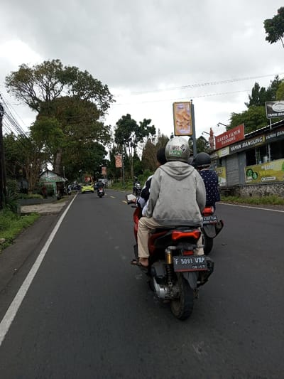 The image depicts a street scene in the city of Taohom, Laos, under an overcast sky. The perspective is from behind a person riding as a passenger on a red scooter, which is moving forward on a dark asphalt road. This passenger wears a grey hooded sweatshirt, light brown pants, and flip-flops, with a light grey helmet adorned with "MISFITS" and a skull logo sticker on the rear. The scooter's license plate, "F 5091 WAP" with "08-27" indicating August 2027, is affixed to the rear. The driver of this scooter wears a black and white long-sleeved shirt, partially visible. Immediately behind, a second person on a dark scooter wears a dark helmet and a dark blue patterned shirt; its license plate "B 4707 YLM" is partially visible.
The dark asphalt road features a solid white line on its left edge. In the midground, a yellow car and additional motorcycles are observed moving or parked. To the right, a building displays an orange sign reading "BAKSO YAMIN BANDUNG" and "YAMIN BANDUNG", with a partially obscured sign below indicating "Bakso". The building's front features partially lowered metal roller shutters. Above the building, a vertical billboard on a pole showcases an image of bread rolls or pastries, with visible text "Olafi ?". The environment is characterized by lush green foliage from large trees and smaller bushes lining both sides of the road, with overhead power lines visible on the left.