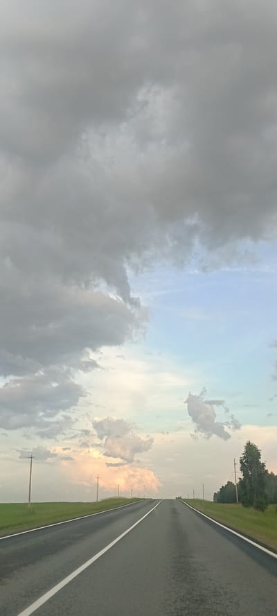 The image depicts a view from a car driving on a straight asphalt road through a rural landscape. The road has a white dividing line. To the left of the road is a green field. To the right of the road, there is also a green field with some trees and power lines visible in the distance. The sky is cloudy, with grey clouds dominating the upper portion of the image and lighter, scattered clouds with hints of pink and yellow near the horizon. The overall impression is that of a drive through the countryside, possibly in the evening or under overcast conditions, in the vicinity of Малышево, Russia. There are no people or animals visible in the image.