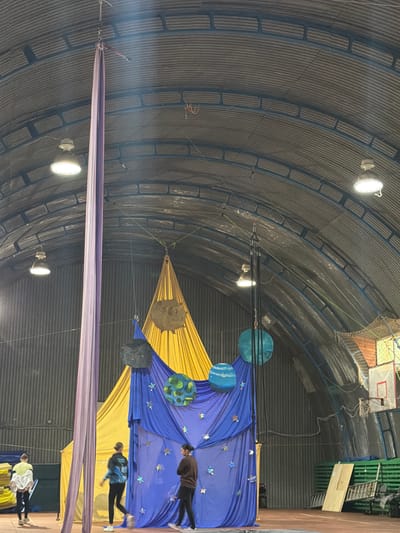 The image depicts an indoor circus or performance space with a high, corrugated metal roof. A long, purple aerial silk hangs from the ceiling on the left side of the frame, alongside a yellow and blue fabric backdrop decorated with stars and planet-like spheres. Two individuals are visible in the foreground, facing away from the camera and looking towards the backdrop. One person, wearing a dark jacket and pants, stands to the right, while another, wearing a blue and black jacket and black pants, is positioned to the left and slightly ahead. A third person in a white jacket and black leggings is partially visible on the far left, walking away from the backdrop. In the background, a basketball hoop is mounted on the wall. The floor appears to be a reddish-brown surface, possibly a sports mat. The overall impression is of a practice or setup for an aerial performance, potentially with a celestial theme.