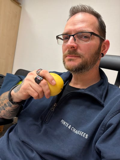A male subject is depicted seated indoors at a facility belonging to the Administration des Ponts et Chaussées in Grevenmacher, Luxembourg. The subject is an adult male with short, dark hair, a trimmed beard and mustache, and dark-rimmed eyeglasses. He has visible ear gauges in both earlobes and tattoos on his left arm and the right side of his neck. He is wearing a navy blue quarter-zip pullover with "PONTS & CHAUSSEES" embroidered in white on the chest, over a black t-shirt. On his left hand, he wears a dark braided leather bracelet and two prominent metal rings; one ring displays a red cross motif, and the other is a black signet ring with a white design. He is holding a single yellow mango fruit in his left hand. The subject is looking directly towards the camera. The background features a plain white wall and a portion of a light-colored wooden cabinet. A black office chair backrest is visible behind the subject's right shoulder.