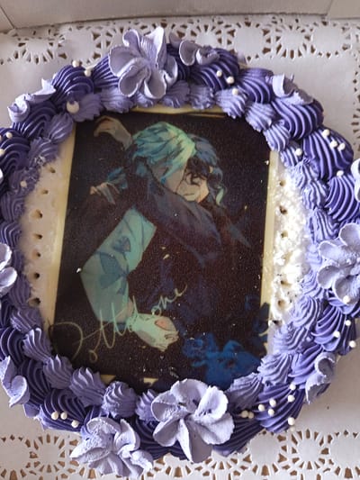 A top-down view of a decorated cake. The cake is round, resting on a white, lacy doily. The center of the cake features a rectangular edible image depicting two characters embracing. The frosting is a light purple color, arranged in swirls and flower shapes around the perimeter of the edible image. Small white sugar pearls adorn the frosting. The location is Novoe Mart'yanovo, Russia.