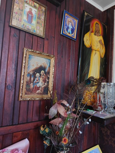 In Kotelva, Ukraine, a wooden-paneled wall serves as a backdrop for several religious icons. A large icon of Mary, with numerous saints surrounding her, hangs prominently. Below it, a smaller, ornately framed icon depicts the Nativity scene. To the right of the Nativity icon, a framed image shows Jesus in a golden robe. The wall is decorated with a vase of dried flowers and peacock feathers, and a crystal vase sits on a shelf. The lighting is dim, suggesting an indoor setting, possibly a home or a place of worship.