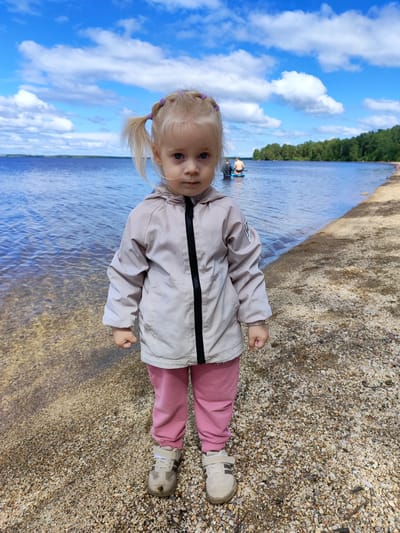 A young Caucasian girl with blonde pigtails is standing on a pebbly shore at the edge of a body of water. She is wearing a light-colored hooded jacket with a black zipper and pink pants, and beige sneakers. She is looking directly at the camera with a serious expression. In the background, the water is blue, reflecting a cloudy sky. Further out on the water, two people are visible on a paddleboard. The shoreline extends to the right, lined with trees in the distance. The overall setting appears to be a natural outdoor environment, possibly a lake or a large river, located in the city of Snezhinsk, Russia.