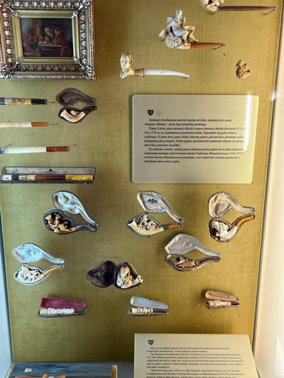 The image shows a museum display of elaborately carved meerschaum pipes, likely in the Trakai Island Castle Museum in Trakai, Lithuania. The display features numerous pipes and pipe cases arranged on a textured, olive-green wall. In the upper left corner, a small framed painting depicts a group of men gathered around a table, possibly in a tavern setting. To the right of the painting, a small, carved bust of a man's head serves as the bowl of a pipe, which extends to the right. Another small carved figure of a cherub is positioned to the right of the bust.
The majority of the display consists of various meerschaum pipes and their associated cases. Many of the pipes are intricately carved with figures, including reclining nudes, mythological scenes, and human busts. Some of the pipes are presented open within their velvet-lined cases, showcasing the detailed craftsmanship. One case in the lower center holds a pipe with multiple carved figures along its length. To the right of that, another case contains a pipe with a carved human figure at the bowl. In the upper right quadrant, a pipe with a seated figure is visible in its case. A rectangular case at the top center contains two pipes.
A descriptive plaque is mounted on the wall below the pipe carvings. The plaque has a heading with "LT" and "EN" indicating multilingual information. The English text describes the creativity in pipe carving using meerschaum, highlighting that the industrial production of foam pipes began in 1750. It mentions Karl Korvach of Hungary as one of the first to carve meerschaum pipes and notes that the pipes were polished with wax to achieve a yellow hue. The text also refers to Karl Friedrich Hieronymus von Münchhausen and his supposed stories told while smoking these pipes. The overall environment appears to be a well-lit museum interior, with the display cases and artifacts well-preserved and presented for public viewing.