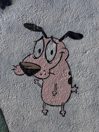 A depiction of the cartoon character "Courage the Cowardly Dog" is painted on a textured, light gray wall in the city of Tomé, Chile. The character, rendered in its characteristic pink hue with black outlines, features large white eyes with black pupils, a dark brown snout, and black floppy ears. A faint smile is visible, accompanied by a slightly raised right front paw. Black spots are present on its body. The coarse texture of the wall is evident through the paint. Strong overhead lighting casts subtle shadows beneath the character's ears and chin. A partial green element is visible at the bottom-left corner, suggesting a larger mural.