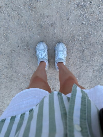 The image, taken from a high angle, shows a person's legs and feet standing on a gravel path in Nîmes, France. The person is wearing white sneakers with white laces and a white and green striped shirt partially visible at the bottom. The ground is composed of small, light-colored gravel and pebbles. The lighting suggests it is daytime.