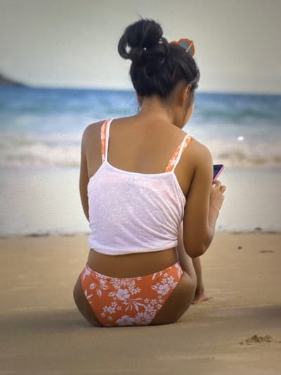 A person is sitting on a sandy beach, facing away from the camera, looking at their phone. The person has dark hair pulled up in a bun and is wearing a white tank top with orange straps and orange floral-patterned bikini bottoms. The ocean and sky are visible in the background. This image was not taken in Bevern, Germany.