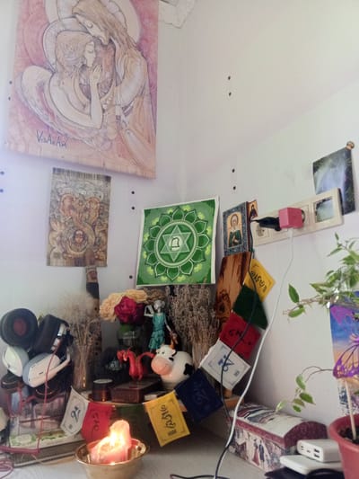The image shows an indoor shrine or altar space, likely in a private residence in Пожарское сельское поселение, Ukraine. The scene is filled with various spiritual and decorative objects. Dominating the upper left is a large, light-colored painting of two figures embracing, with a signature at the bottom reading "VedAnAM." Below this painting hangs a smaller tapestry with an intricate depiction of an elephant and a child. To the right of these, a green square cloth displays a star-like symbol.
On the surface below the wall hangings, several items are arranged. These include a small statue of a winged female figure, a white cow figurine, and a red dragon figurine. There are also several dried flower arrangements and what appear to be prayer flags with Tibetan script, hanging from a wooden support. A lit candle in a brass bowl is centrally positioned in the foreground, casting a warm glow. To the left, a stack of headphones is visible.
Further to the right, an electrical outlet strip is mounted on the wall with several plugs and a pink charging device. A small potted plant with green leaves and a butterfly decoration is to the far right. The overall impression is of a personal and spiritual space, decorated with a mix of art, figurines, and symbolic items. The lighting appears to be a mix of natural and artificial light, casting soft shadows. The background is a plain white wall, suggesting an interior room.