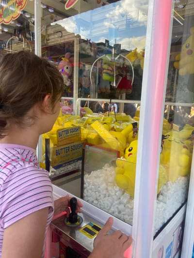 A child is playing a claw machine game. The child, facing away from the camera, is reaching out a hand towards the joystick of the machine. The machine is filled with yellow plush toys, some of which appear to be Pikachus. There are also other toys visible, including a pink character with a bow. The claw machine has a bright pink light strip across the top. The reflection in the glass shows a cloudy sky and buildings. Visible text on the machine includes "BUTTER SQUISHY," "Super Sofet! Slow Rising," "Looks like Butter-Feels Like Squishy," and "NOT REAL FOOD/TOY." There are also labels for "CREDITS," "MONEY," and "TIME." The setting appears to be an arcade or amusement area. The child's focused expression is suggested by their posture. The lighting suggests it could be daytime.