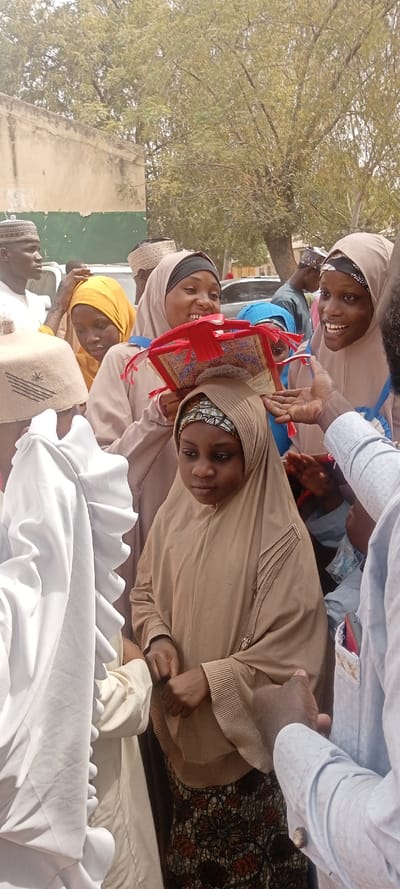 The image captures an outdoor gathering in Sani Mai Nagge, Nigeria, under daylight. In the foreground, a young female, approximately 10-14 years old, stands centrally, clad in a full-length light brown hijab and an embellished brown jilbab featuring decorative pleats and beading on the left side. She wears a patterned undercap and holds her hands clasped, displaying a neutral expression.
To her immediate right, a dark-skinned male hand, wearing a light-blue patterned long-sleeved shirt with cuff links, extends towards her. Behind the young female, two young adult females are positioned, both smiling. The female on the viewer's left wears a light beige hijab with a black undercap and holds a small, ornate red book, likely a Quran, resting on a red decorative stand with tassels. The female on the viewer's right wears a similar light beige hijab with a patterned undercap and a blue ribbon accessory, looking towards the extended hand. A third female in a yellow hijab is partially visible between them.
In the left foreground, the back of an individual wearing a light beige prayer cap with a distinct pattern and a white, ruffled garment is visible. Other individuals, including a male in a white prayer cap on the far left, are present in the mid-ground.
The background features a light-colored building with a green lower section, and multiple mature trees with green foliage. A light-colored vehicle is partially discernible in the left mid-ground. The scene suggests a communal event, possibly a religious ceremony or presentation.