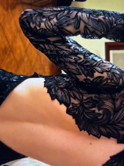A close-up image shows the back and left arm of a person wearing a dark, ornate, long-sleeved gown. The back is partially exposed. The gown has a lace-like pattern of swirling leaves, possibly embroidered or appliqued onto a sheer fabric. Tiny reflective sequins embellish the pattern, adding sparkle. The background is softly blurred, revealing a glimpse of wooden flooring and a framed picture.