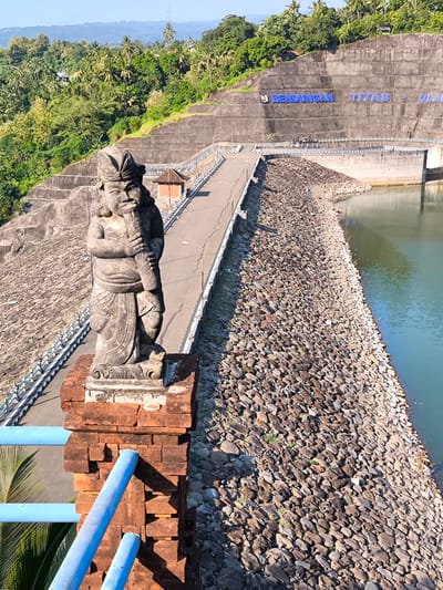 The image captures a detailed view of the Bendungan Titab-Ularan (Titab-Ularan Dam) located near Seririt, Buleleng Regency, Bali, Indonesia. In the immediate foreground, a weathered stone statue of a humanoid figure, sculpted in a traditional Balinese style, stands on a reddish-brown brick pedestal. The figure appears stout, with prominent facial features and stylized hair or headwear, and is holding a long, pipe-like object vertically in its hands. Blue painted metal railings are visible to the left and below the statue, indicating a vantage point from a bridge or walkway.
The midground is dominated by the massive concrete structure of the dam. A paved road stretches across the top of the dam wall, bordered by white railings and streetlights. Prominently displayed on the grey concrete face of the dam, large blue three-dimensional letters spell out "BENDUNGAN TITAB - ULA". A body of green-tinged water, likely the reservoir, is visible to the right of the road, with a steep embankment of large, irregularly shaped rocks forming its edge. Beyond the dam, the terrain rises into lush green hills densely covered with tropical vegetation, including numerous palm trees. Distant mountains or hills are visible under a clear, bright blue sky. The scene is illuminated by strong sunlight, casting distinct shadows, indicative of daytime. No human figures are explicitly visible in the frame.