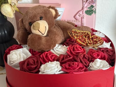 The scene is an indoor setting in Vellmar, Germany, depicting a still life arrangement. A brown plush teddy bear with black bead eyes, a tan snout, and an embroidered black smile is positioned on its left side, resting its head on its left paw. It lies within a red, heart-shaped box. The box is filled with numerous artificial foam roses in shades of dark red and white, many exhibiting a subtle glittery texture. A gold-colored, filigree butterfly ornament is placed among the roses to the right of the teddy bear. In the upper left background, a black, vertically ribbed vase holds dried, light-colored botanical elements. To the upper right background, a stack of light pink gift boxes is visible; one box features a white ribbon with repeating 'CH' logos and green leafy designs, with partially visible text 'HERRERA' below the ribbon. The overall background is a light, neutral wall.