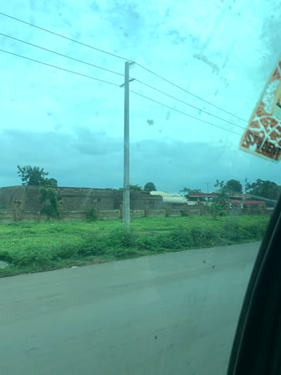 The image depicts a scene in Laminga, Nigeria, characterized by a cloudy sky and a landscape dominated by a long, low-lying building constructed of grey bricks, surrounded by a low stone wall. Lush green vegetation, including grass and trees, flanks the structure and extends towards a paved road in the foreground. A tall utility pole with multiple wires stands prominently near the center of the frame. In the upper right corner, a sticker with the word "STUDENT" is visible, adhered to what appears to be a car window, suggesting the photo was taken from inside a moving vehicle. The overall impression is that of an industrial or institutional complex situated in a semi-rural or suburban area.
