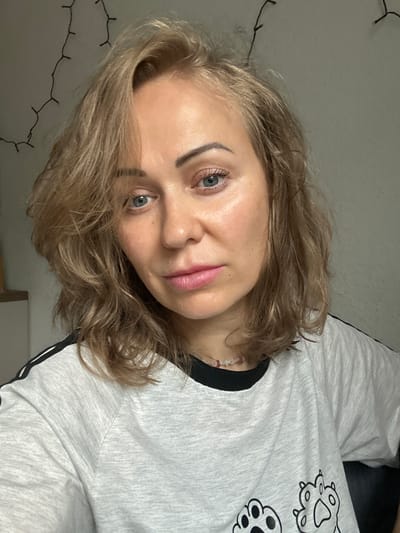 The image shows a close-up, head-and-shoulders shot of a woman with light brown, wavy hair and blue eyes. She is looking directly at the camera with a neutral expression. She is wearing a light gray t-shirt with black trim around the collar and black stripes on the sleeves. A delicate beaded necklace is visible around her neck. The background is a plain, light gray wall with a string of small lights draped across the top left corner. The lighting appears to be artificial and somewhat dim. The scene suggests an indoor setting, possibly a home environment, with no clear indicators of a specific urban location. There is no information available to suggest the image was taken in Velbert, Germany.