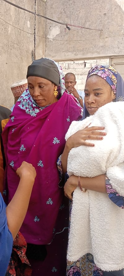 The scene depicts an outdoor gathering in Dan Maliki, Nigeria, set against a weathered, light-colored concrete or stucco wall with visible wires strung across its upper section.
In the foreground, two adult females are prominent. On the left, an adult female with medium-brown skin wears a dark grey head covering over a patterned underlayer and a vibrant magenta fabric draped over her shoulders and torso, adorned with light blue floral embroidery and silver sequins. She wears long, gold, leaf-shaped dangle earrings and looks downwards. Her left side is partially obscured by an outstretched forearm in a blue sleeve, with a hand positioned near her midsection.
On the right, a younger adult female with light-medium brown skin wears a patterned headscarf (blue, purple, pink, yellow motifs) and a dark blue garment. She holds a white, fluffy, sherpa-like blanket or garment across her front with both hands, her fingernails painted red. A copper-toned, link-style bracelet is visible on her right wrist. She has a small nose piercing on her left nostril and looks slightly towards the viewer.
In the background, behind the two women, an adult male with a shaved head is visible, wearing a light-colored shirt and a watch on his left wrist. His right hand is raised to his chin. Further to the left and behind the first woman, a portion of another male individual is visible, wearing a light-colored, patterned cap or kufi. Some exposed brickwork is discernible in the distant background.