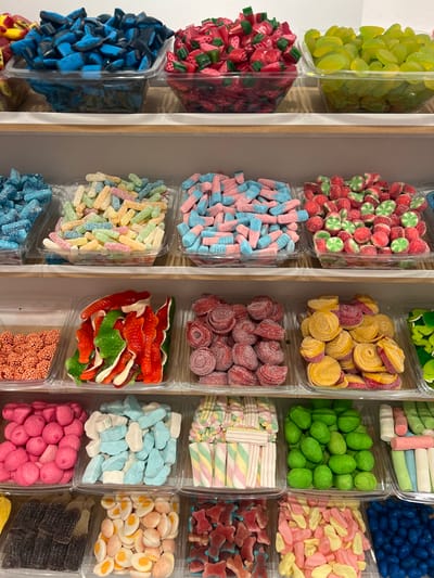 A brightly lit candy store in Port De Pollença, Spain, displays a wide assortment of colorful gummy candies arranged in clear plastic containers on tiered shelving. The candies are organized by type and color, creating a visually appealing mosaic. Visible candy types include gummy watermelon slices, green grapes, sour belts in various fruit flavors, gummy sharks in red and green, swirled fruit candies, gummy marshmallows, sour balls in green, and gummy bears in red. The store appears clean and well-stocked, with the focus solely on the candy displays. There are no people or other objects visible in the scene.