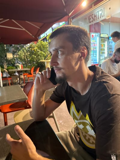 A man, likely in his late 20s or early 30s, with short dark hair, a beard, and dark eyes, is seated at an outdoor cafe table, speaking on a dark smartphone held to his right ear. He is wearing a dark t-shirt with a yellow and white circular logo on it, and a silver chain around his neck. His left hand is resting on the table, palm up. In the background, other cafe tables and chairs are visible, as is a building with "MJESHTRI" signage. The setting appears to be a street-level establishment in Bashkia Durrës, Albania, possibly a restaurant or cafe, with outdoor seating and illuminated by artificial lighting. The time appears to be evening or night.