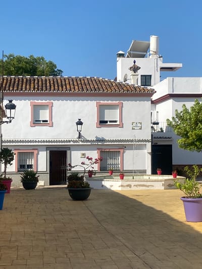 The image shows a bright, sunny day in the town of Cancelada, Spain. A white building with terracotta roof tiles and pink trim around the windows is the primary focus. A small fountain is in front of the building, surrounded by potted plants. The ground is paved with light-colored rectangular stones. Trees and other buildings are visible in the background. The scene appears tranquil and well-maintained, suggesting a peaceful residential area.
