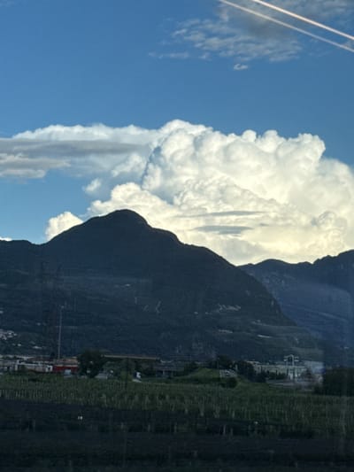 This is a vertical, medium-angle, outdoor shot of a mountainous landscape under a blue sky with clouds, and a village at the base of the mountains. The sky is mostly clear blue, with a few wispy white clouds scattered across the top. Towards the center of the frame, a large, puffy white cumulus cloud formation dominates the sky. In the middle ground, a dark, silhouetted mountain range stretches across the width of the image. The foreground shows a strip of dark green vegetation, possibly vineyards, followed by a village with buildings of various sizes and colors. Some structures are white, with one building featuring a distinct white archway. Power lines and utility poles are visible against the dark mountain slopes. The scene appears to be captured during daylight, likely late afternoon or early evening due to the lighting. The location is identified as Lavis, Italy.