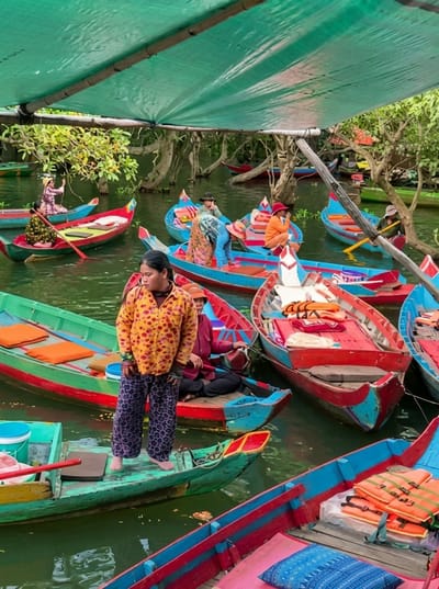 This is a photograph taken in the city of Soutr Nikom, Cambodia, featuring a collection of colorful boats on a body of water. A large green tarp, supported by wooden poles and a beam, stretches across the top of the frame, providing shade. In the foreground, a woman wearing a yellow floral-patterned shirt and patterned purple pants stands on a green boat, facing left. Behind her, a person in a maroon shirt is seated in a red and blue boat. Several other boats are visible, many adorned with vibrant red, blue, and green paint. Some boats contain passengers dressed in colorful clothing, including women in conical hats. In some boats, life vests are neatly stacked. The water is murky green, and the surrounding environment is characterized by trees with visible branches and foliage, suggesting a natural or semi-natural setting. The overall scene depicts a bustling water market or a tourist activity involving boat transportation.