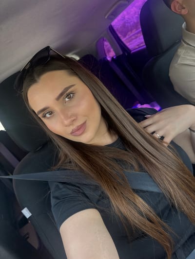 A woman with long, brown hair and light skin is taking a selfie in the passenger seat of a car. She is wearing a black t-shirt and a seatbelt. Sunglasses are resting on top of her head. Her left hand, adorned with a ring, is resting on her chest. A second person, whose face is not visible, is seated in the driver's seat, wearing a light-colored collared shirt. The car's interior is dark, with purple ambient lighting visible through the windows, suggesting it is nighttime or dusk, possibly in the vicinity of Razliv, Bulgaria. The scene appears to be a casual moment captured during a car ride.