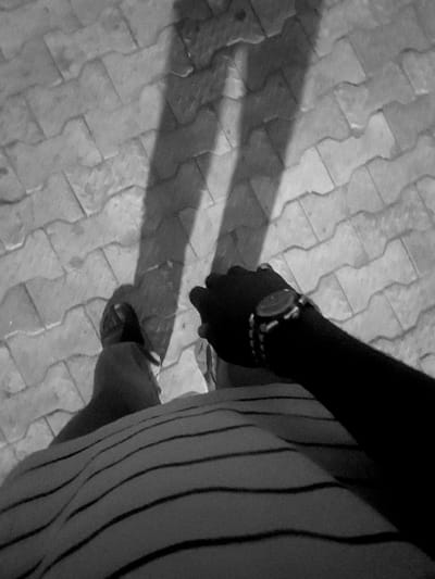From a high angle, a person stands on a patterned tile surface. A dark shadow stretches across the tiles. The person's arm, adorned with a watch, is visible on the right side, along with part of their striped garment. The person is wearing sandals on their feet. The scene is monochromatic and emphasizes the contrast between light and shadow on the textured ground.