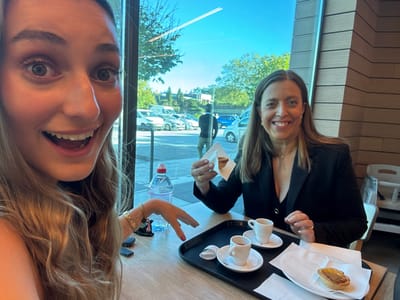 Two females are seated at a light-colored wooden table inside a cafe with a large window overlooking an urban outdoor scene in Leiria, Portugal. The female in the foreground, positioned left, has long blonde hair, wide eyes, and an open-mouthed smile, facing the camera as if taking a selfie. Her right hand, featuring red nail polish and a gold bracelet, rests on the table. The female in the background, right, has long brown hair and a smiling expression, holding a pastel de nata wrapped in a paper napkin in her left hand. Her right hand is on the table. A black serving tray on the table contains two white espresso cups on saucers, each holding coffee, and a third pastel de nata resting on a white paper napkin. A clear plastic water bottle with a red cap is also on the table. Through the window, a bright blue sky with a faint contrail is visible above green trees and a paved area with several parked vehicles. A male figure is partially visible walking in the mid-ground, and a blue car is further in the distance. A white high chair is present in the far right background of the interior.