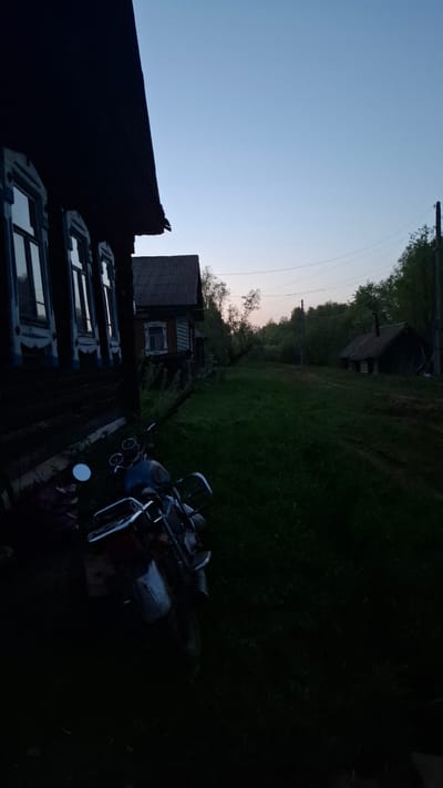 The image depicts a rural scene in the city of Шешурга, Russia, likely at dusk or dawn. A dark wooden building with visible windows occupies the lower left. In the foreground, a lawn mower rests on the grassy ground. In the background, a smaller wooden structure is visible amidst trees and vegetation. Power lines stretch across the upper part of the image against the light blue sky. The scene is dimly lit, with shadows obscuring many details.