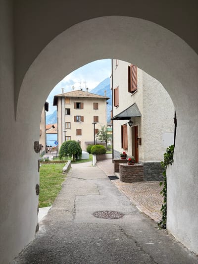 The image captures a view through a curved archway, revealing a street scene in Molveno, Italy. The perspective is from inside an archway, with its textured, pale exterior dominating the foreground and left side of the frame. Through the opening, a narrow paved street or alleyway is visible, flanked by buildings with light-colored stucco exteriors and dark wooden shutters. Several windows are open, revealing the interiors of the buildings.

To the right of the alley, a patch of green grass slopes upwards, bordered by a low concrete wall. Further up the alley, a circular cobblestone pattern is embedded in a small wall or planter. A few small potted plants with red flowers are visible near a doorway. Green ivy climbs on a section of the wall to the right of the alley.

In the distance, at the end of the alley, a street sign with a blue circle and a red bar is visible, indicating a traffic regulation. Beyond this, a glimpse of mountainous terrain under a cloudy sky can be seen. The sky suggests it is daytime, likely overcast. There are no people visible in the scene. The overall impression is one of a quiet, European village setting. The photograph is oriented vertically.