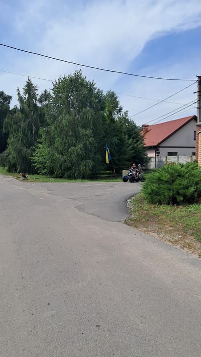 In Kryvushi, Ukraine, a two-lane asphalt road curves to the right, bordered by grass and trees. On the left side of the road, a person is lying on the ground near a patch of grass. On the right side of the road, two people are riding an ATV. Behind them, a Ukrainian flag is flying from a pole. A residential building with a red roof and a brick chimney is visible in the background, along with a section of a brick wall and a dense green hedge. The sky above is a mix of blue with scattered white clouds. Power lines are visible overhead.