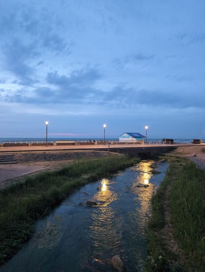 The image depicts a tranquil evening scene in Veules-les-Roses, France. A small, clear stream flows through the foreground, its surface reflecting the warm glow of streetlights. Lush green grass lines the banks of the stream. In the background, a promenade runs alongside a body of water, possibly the English Channel. A few scattered streetlights illuminate the promenade, and a small, blue and white building with text "VIE À LA PLAGE" is visible. The sky is a deep blue with scattered clouds, suggesting either dusk or early dawn. A few indistinct figures can be seen on the promenade in the distance. The overall atmosphere is peaceful and serene, capturing the quiet beauty of the seaside town.