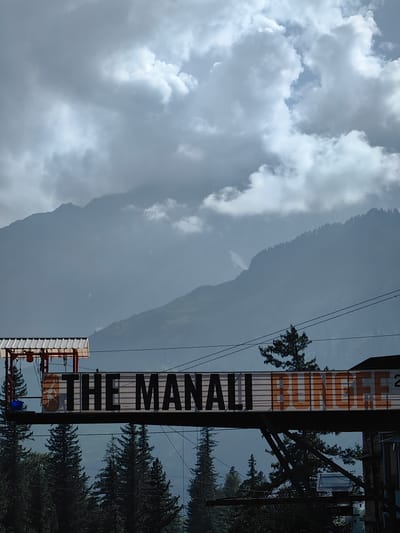 The image depicts the "The Manali Bungee" sign at a bungee jumping site in Kothi, India. The sign, written in large black and orange letters, is mounted on a horizontal structure spanning across a scenic mountain landscape. Dark, overcast clouds partially obscure the rugged, mist-covered mountain peaks in the background. In the foreground, lush green pine trees are visible, framing the bungee platform. A blue bucket hangs from the left side of the platform. The overall atmosphere is dramatic and imposing, with the weather adding to the sense of adventure and natural grandeur of the location.