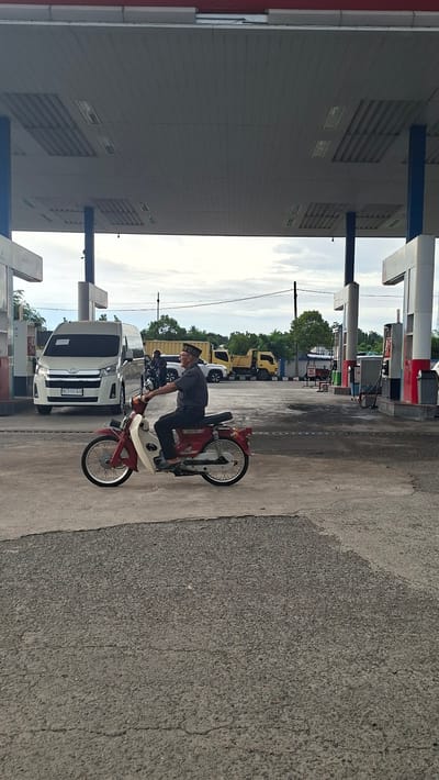 At a gas station in Keuniree, Indonesia, an elderly man wearing a dark shirt and a patterned cap rides a red and white motorcycle. The man is positioned in the foreground, facing right, with his hands on the handlebars. In the background, a white van with a license plate displaying "BL 1137 AZB" is parked. Other vehicles, including yellow trucks, are also present at the gas station. The scene takes place under a covered roof with lighting fixtures. The ground is paved with asphalt. The sky above is cloudy.