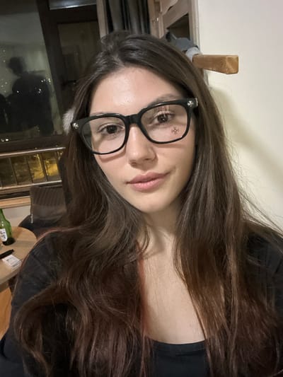 A female subject, appearing to be in her twenties or early thirties, is positioned centrally, facing forward and looking directly at the viewer. She has long, dark brown hair with subtle reddish undertones, worn down and wavy. She is wearing black-framed glasses featuring decorative cross-like emblems on the outer corners. Her expression is neutral with a slight smile. She is dressed in a dark-colored, long-sleeved top with a wide neckline. A small, white, possibly fluffy object is partially visible near her left temple.
The scene is an indoor, likely residential, setting within Huangdao District, China. To the left of the subject, a window reveals a dark exterior, indicating night, with blurred distant lights. A faint, dark reflection of a person, possibly male and wearing a hat or hood, is discernible in the window. Below the window, a small, light-colored round table is present, holding a green glass bottle (consistent with a soju or beer bottle), a dark rectangular object, and a stack of small white rectangular items. Part of a dark-colored upholstered chair or sofa is visible near the table. To the right of the subject, the background consists of a plain, light-colored wall. A wooden beam or protrusion extends from the upper right portion of this wall, with a dark, indistinct object hanging or resting above it.